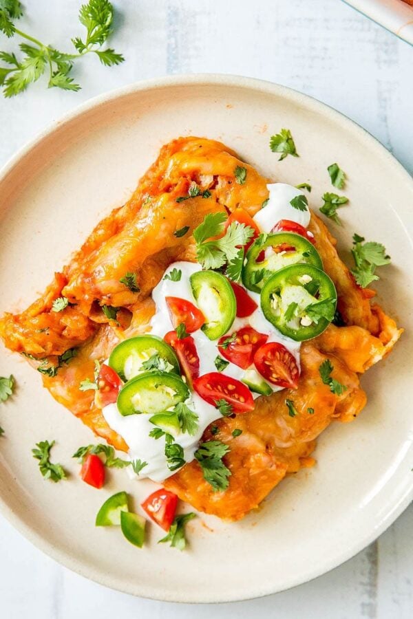 Homemade cheese enchiladas on a white plate topped with sour cream, jalapeños, tomatoes and cilantro.