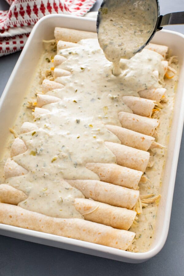 Creamy white sauce is being poured over rolled, unbaked enchiladas.