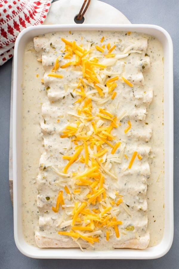 Rolled chicken enchiladas are topped with white enchilada sauce and shredded cheese.