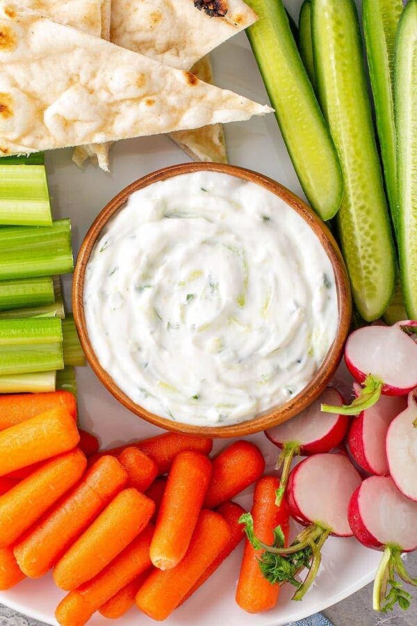 A close up of a wooden bowl of Tzatziki Sauce surrounded by carrots, celery, pita chips and cucumber spears