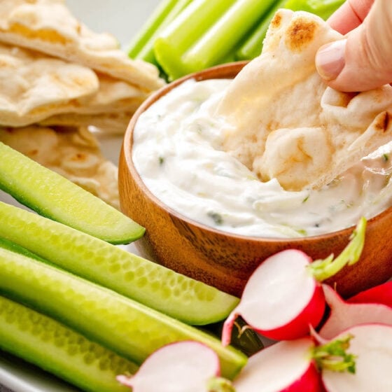 A pita chip is being dipped into a bowl of Tzatziki that is surrounded by vegetables