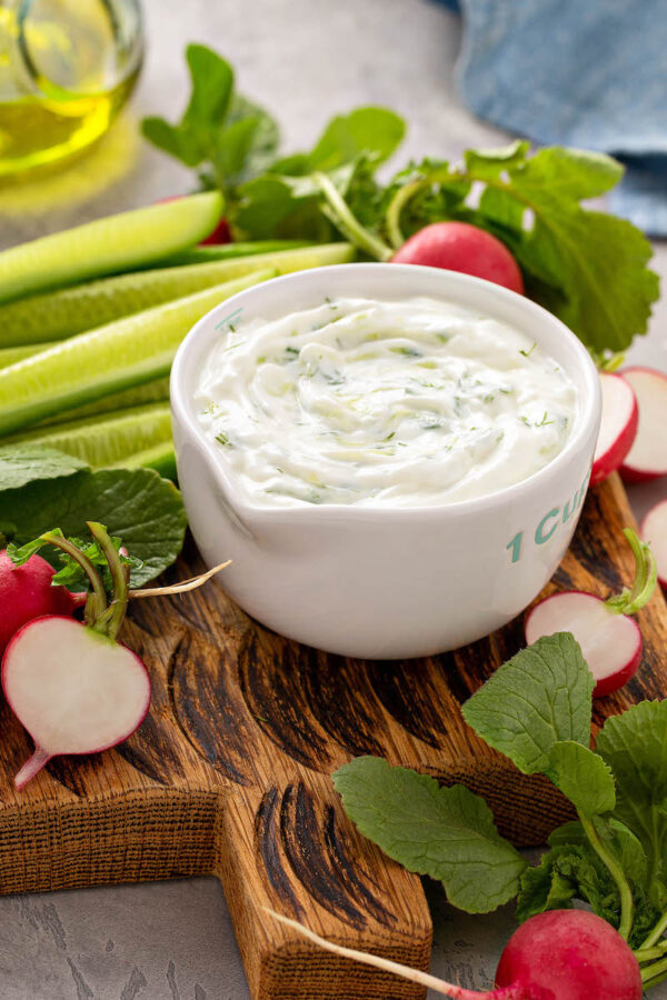 A small bowl of Greek cucumber dip on a wooden board surrounded by radishes and cucumber spears