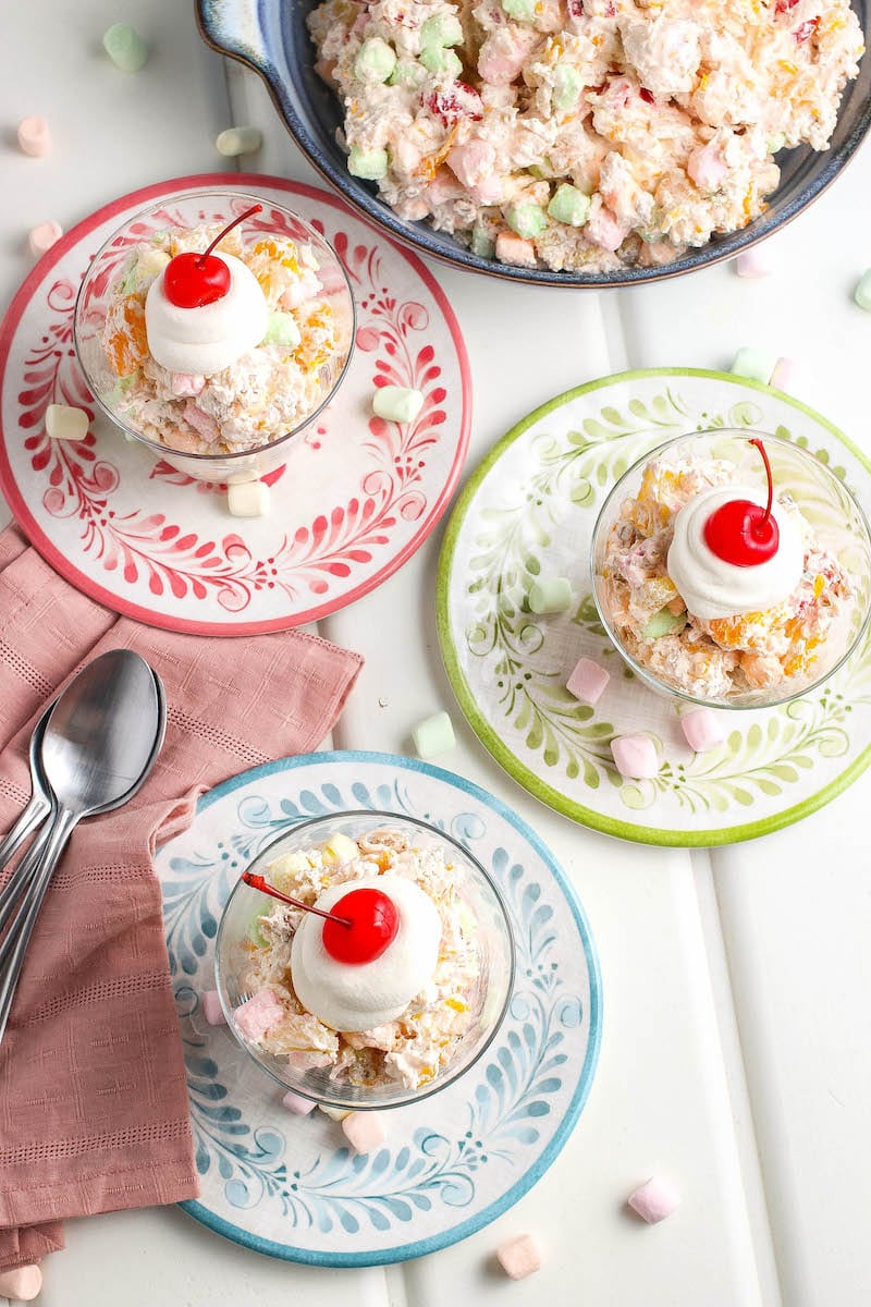 3 servings of Ambrosia salad on colorful saucers topped with whipped topping and a cherry. 