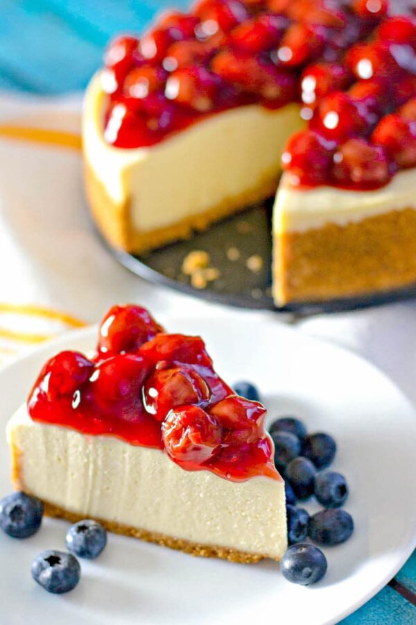 A perfect slice of cheesecake one a white plate surrounded by blue berries. 