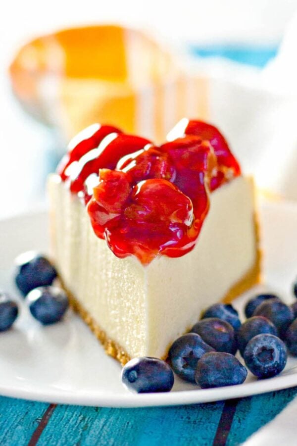 A perfect slice of cheesecake one a white plate surrounded by blue berries. 