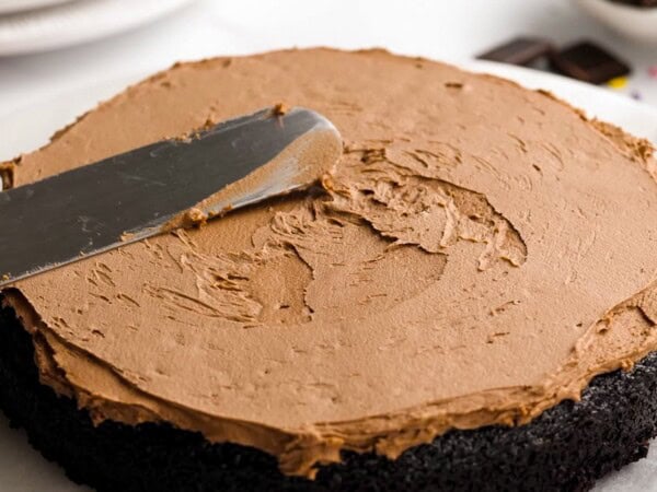 Rich chocolate frosting is being spread over a layer of chocolate cake. 