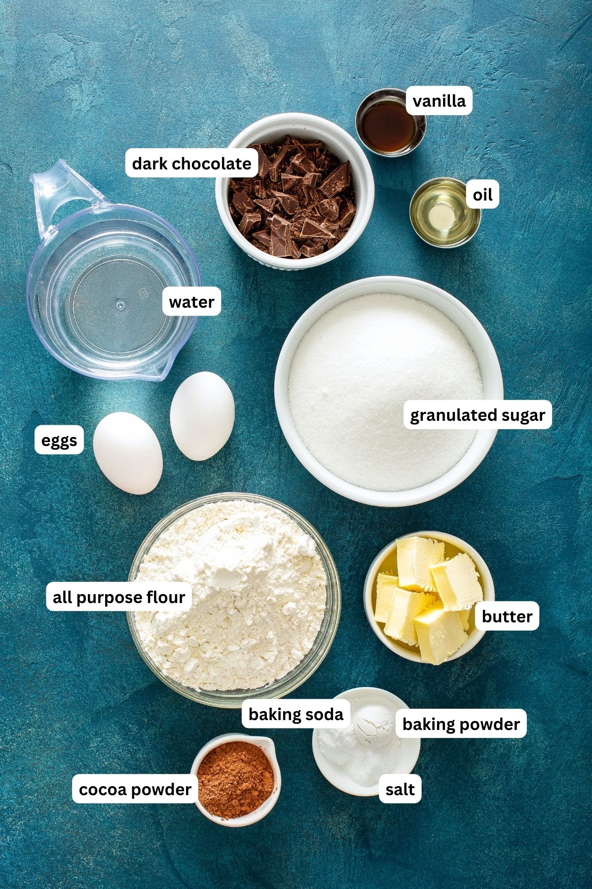 The ingredients for chocolate cake recipe in order from top to bottom: water, dark chocolate, vanilla, oil, eggs, granulated sugar, all purpose flour, butter, cocoa powder, baking soda, baking powder, salt. 