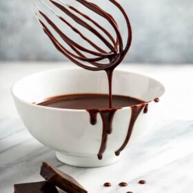 Chocolate sauce in a white bowl with a whisk and chocolate bar on marble.