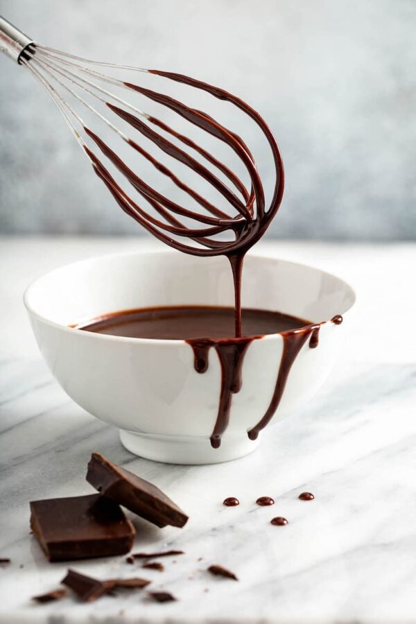 Chocolate sauce in a white bowl with a whisk and chocolate bar on marble. 
