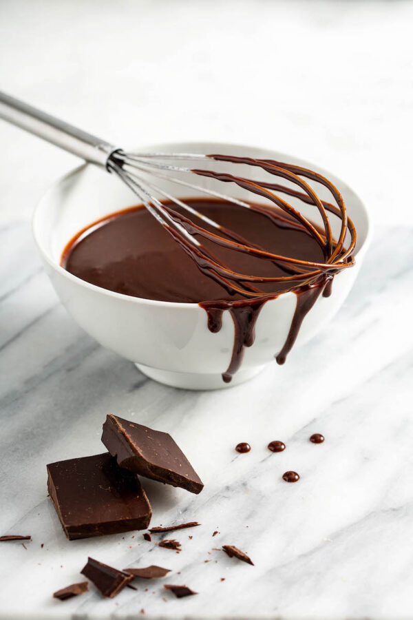 Chocolate sauce on a whisk in a bowl with chocolate squares. 