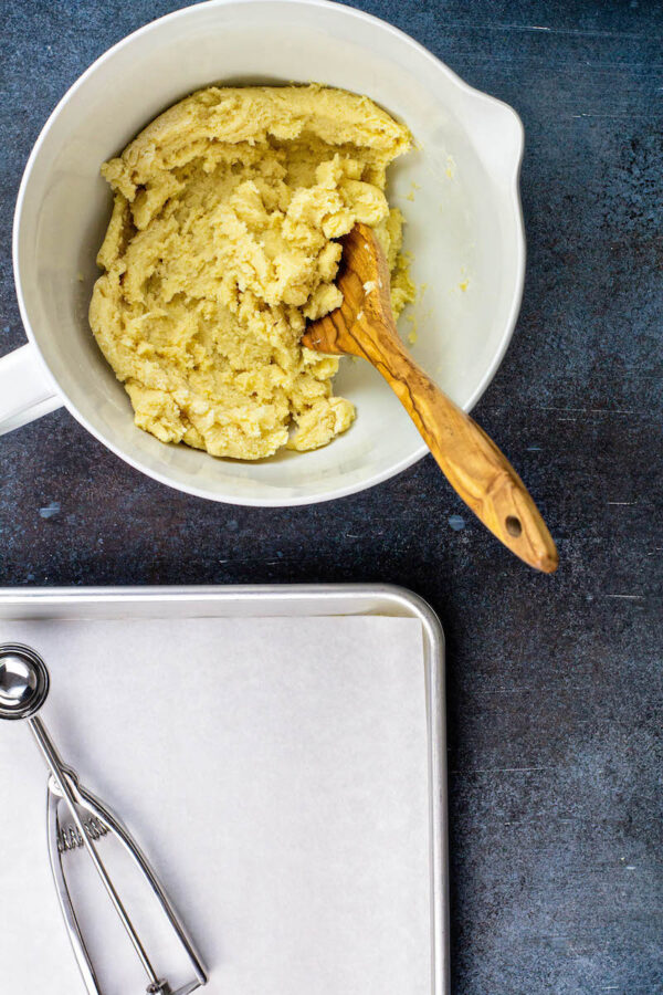 Meltaway cookie dough in a white bowl with a spoon and a baking sheet with a cookie scoop. 