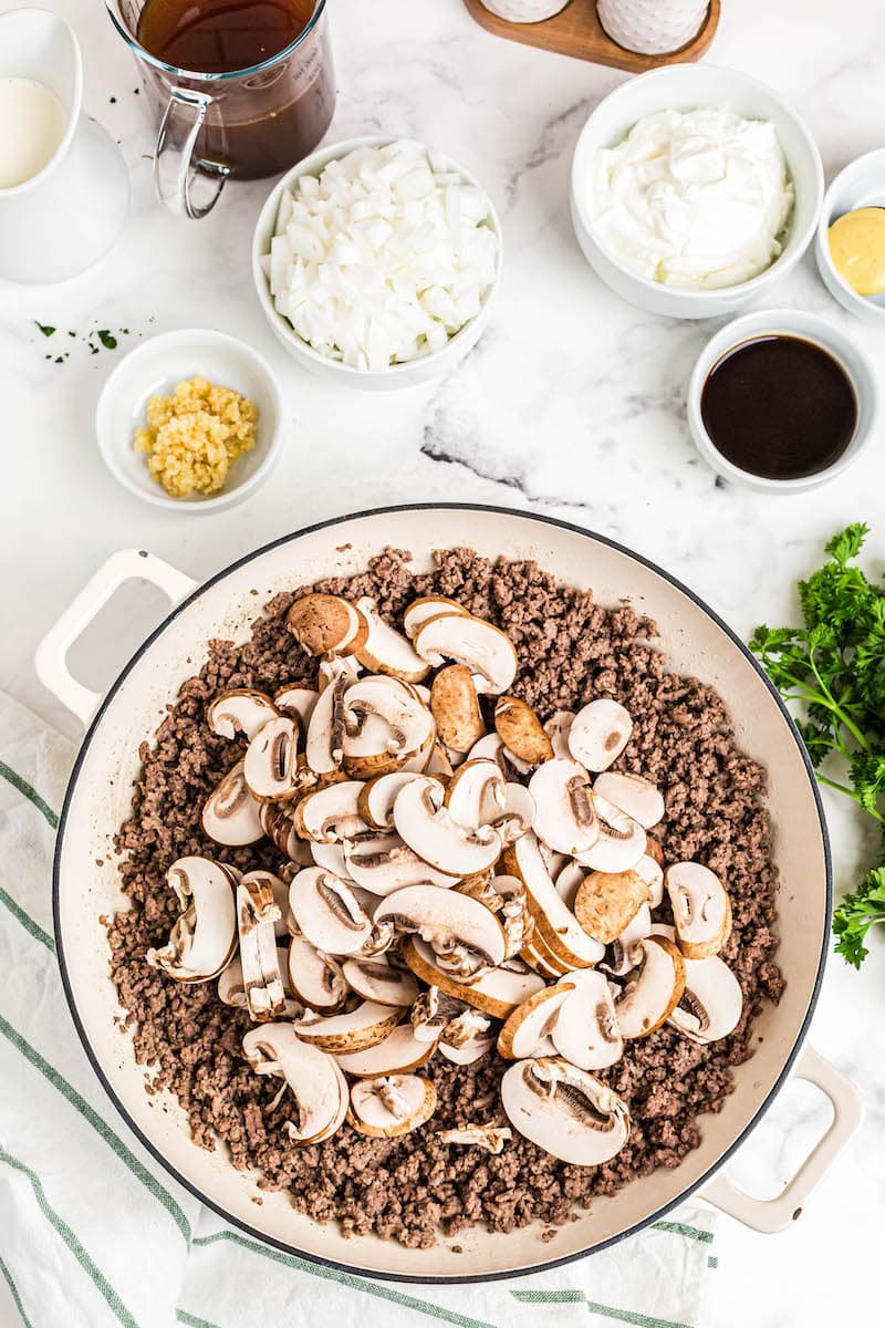 Large pan filled with ground beef and mushrooms with other ingredients around.