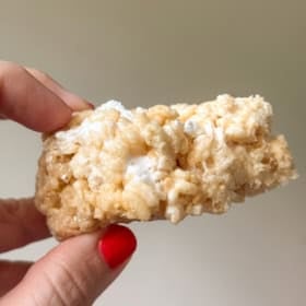 A hand with red nail polish holds a thick, gooey Rice Krispies treat with visible melted marshmallows against a plain background.