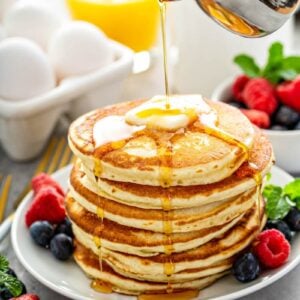 Stack of fluffy pancakes on a white plate with fresh fruit and syrup being poured on top of them.
