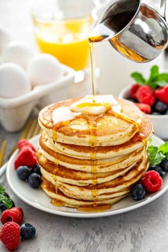 Stack of fluffy pancakes on a white plate with fresh fruit and syrup being poured on top of them.