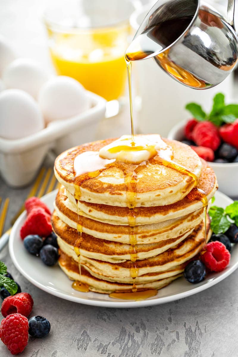 Stack of fluffy pancakes on a white plate with fresh fruit and syrup being poured on top of them. 