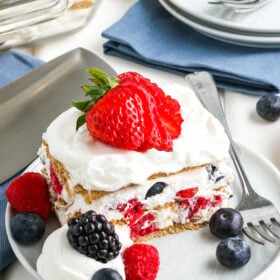Slice of icebox cake on a plate with a sliced strawberry on top, more berries on the plate, and a fork next to it.