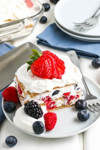 Slice of icebox cake on a plate with a sliced strawberry on top, more berries on the plate, and a fork next to it.