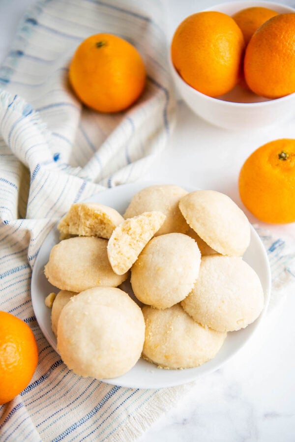 Orange whipped shortbread cookies on a white plate with one torn in half to see the inside.