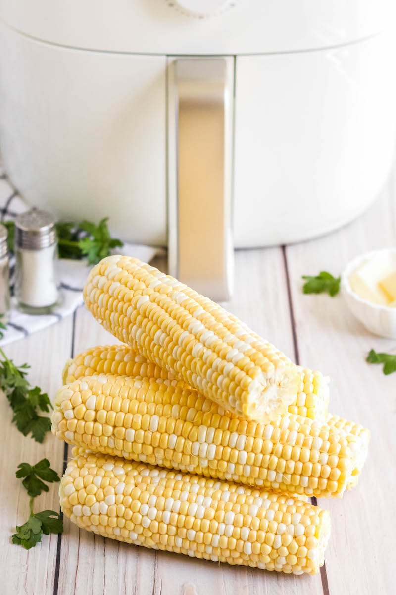 4 stacked corn cobs without seasoning on a table in front of the air fryer.
