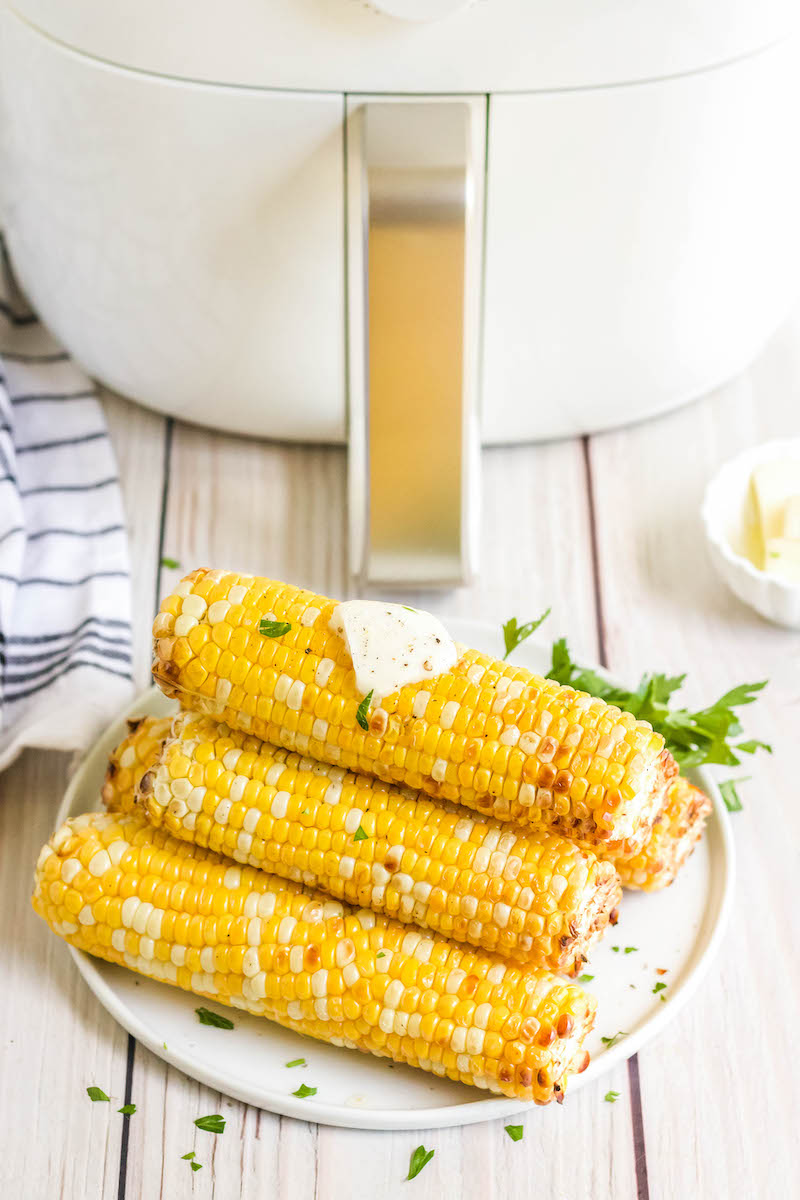 Corn on the cob on a white plate with butter on top with an air fryer in the background.