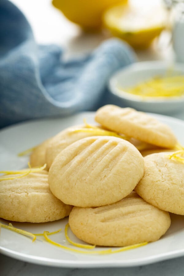 Lemon whipped shortbread cookies on a white plate with lemon zest and a blue tea towel.