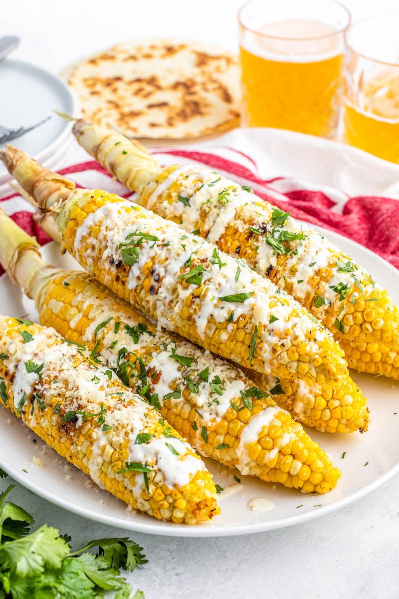 6 corn cobs on a plate with sour cream sauce, cotija cheese, and fresh cilantro.
