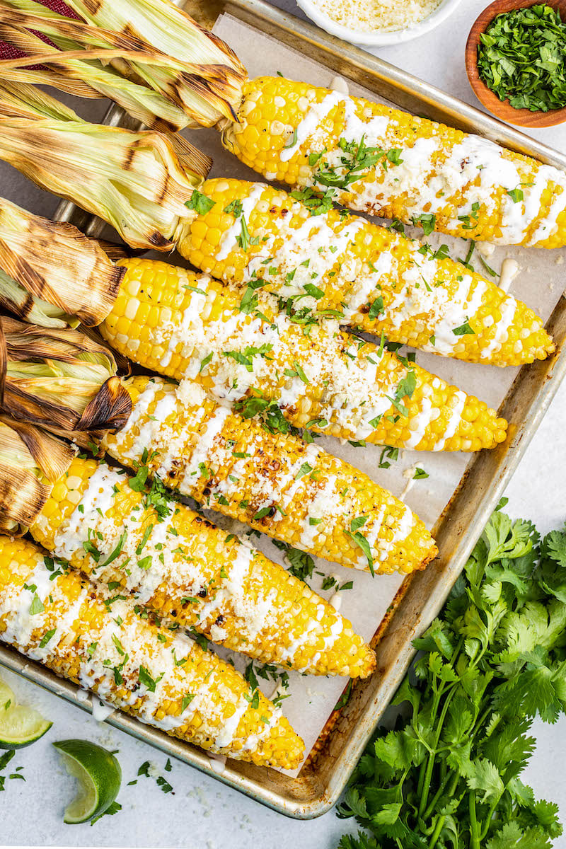6 grilled corn cobs with the husks peeled back on a pan, with sour cream sauce on the top of the cobs.
