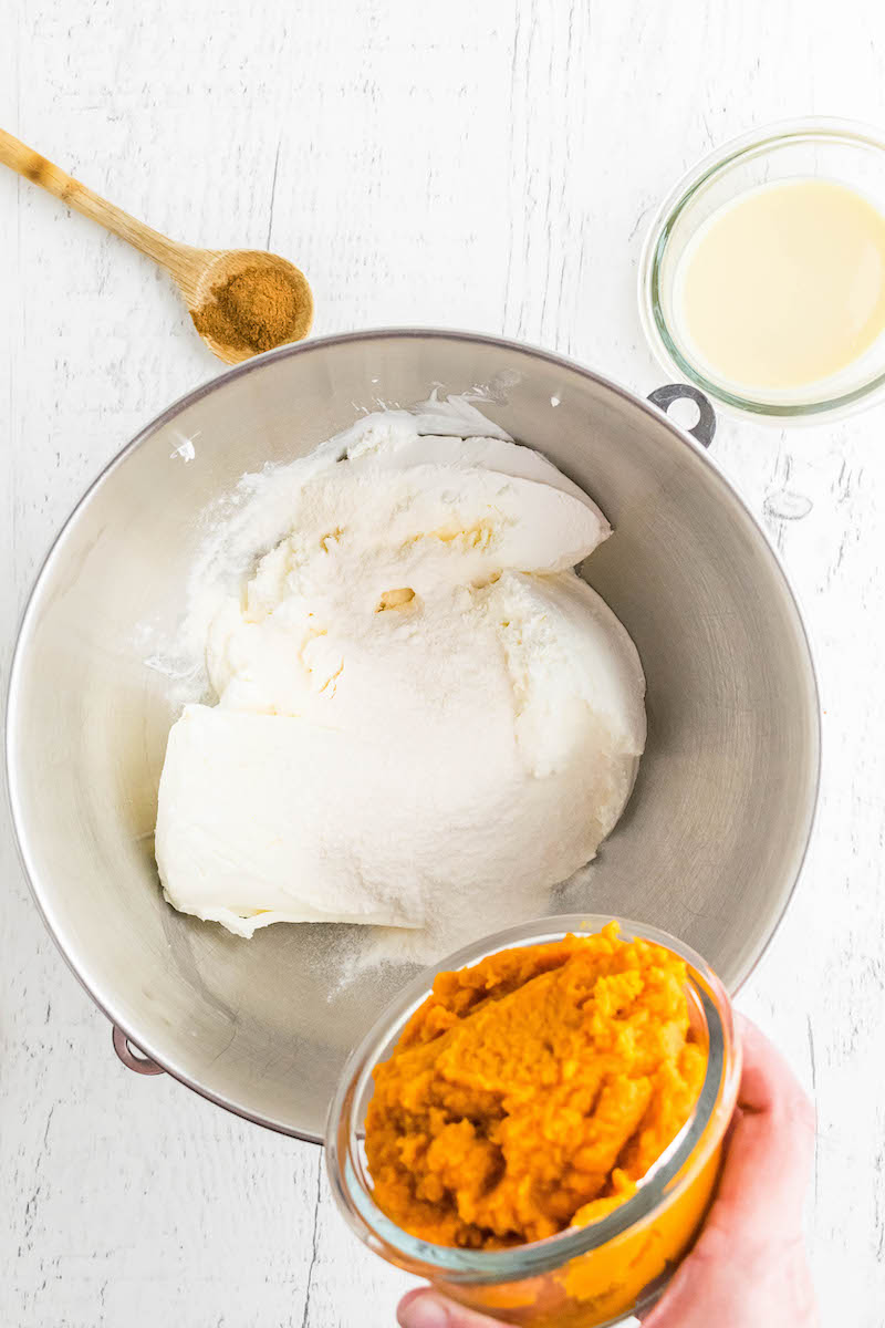 Mixing bowl with instant pudding and cream cheese mixture in it, with a bowl of pumpkin puree being poured into it.