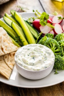 Tzatziki sauce in a bowl next to pita bread, cucumbers, and other vegetables.