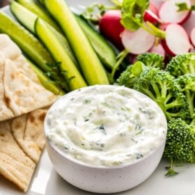 Tzatziki sauce in a bowl next to pita bread, cucumbers, and other vegetables.