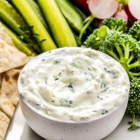 Bowl of cucumber dip with veggies on a plate.