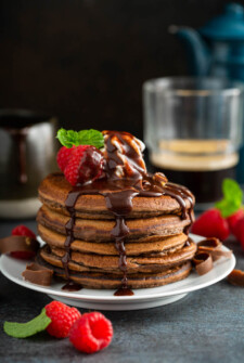 Chocolate pancakes stacked on a white plate with chocolate sauce and raspberries on top.