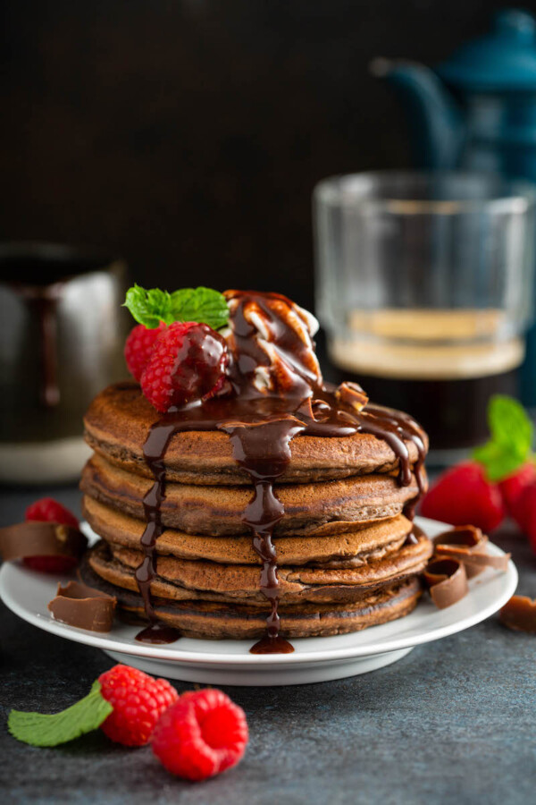 Chocolate pancakes stacked on a white plate with chocolate sauce and raspberries on top.