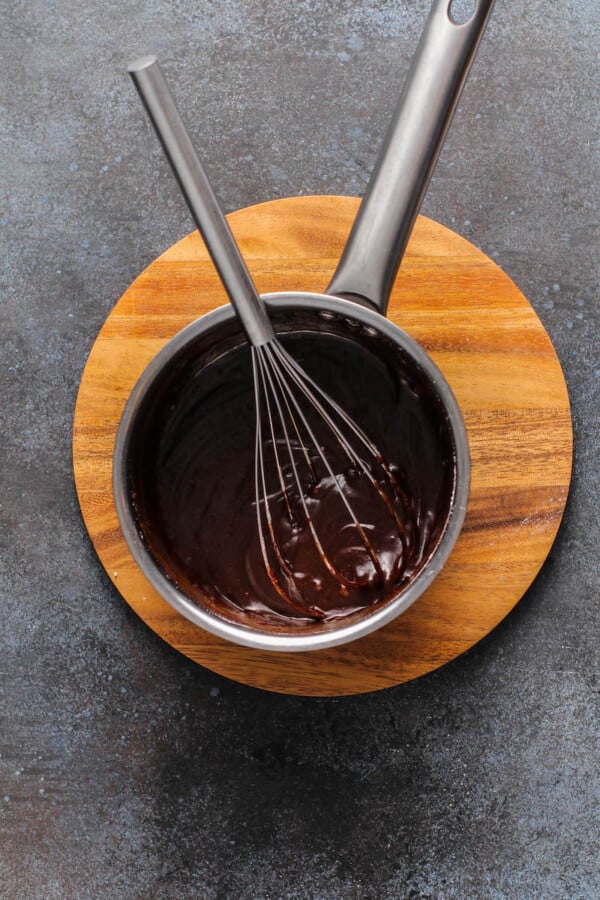 Chocolate sauce in a sauce pan with a whisk inside on a wood cutting board.