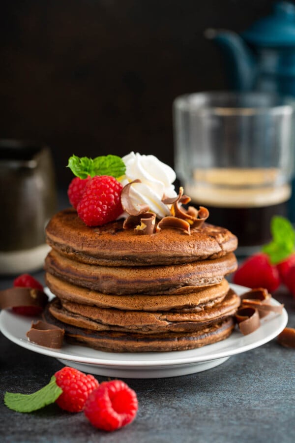 Fluffy chocolate pancakes on a white plate with whip cream and raspberries on top and on the side.