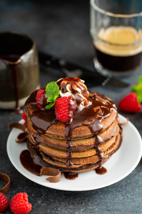 Chocolate pancakes with chocolate sauce and raspberries on top with a cup of coffee in background.