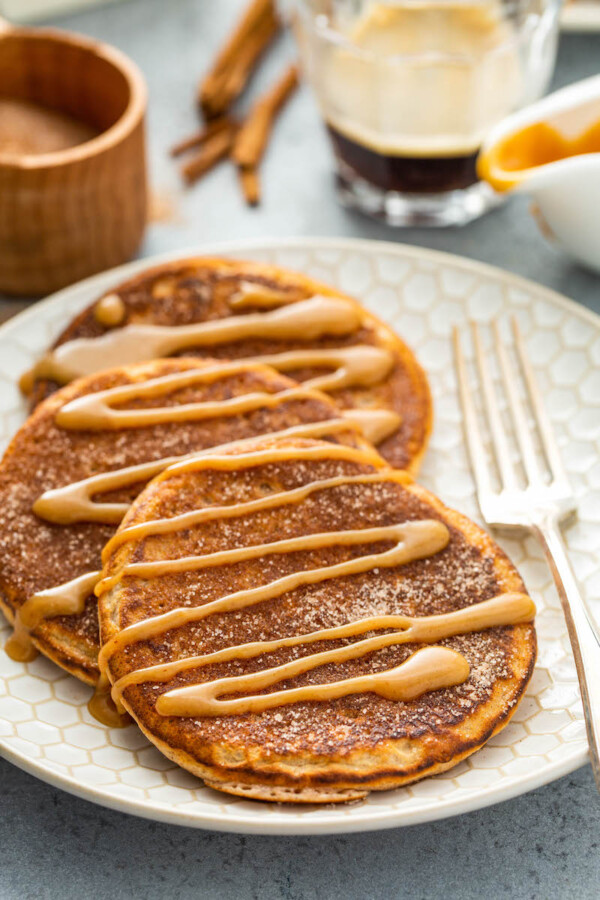 Three churro pancakes ona plate with a fork and cinnamon sugar in a bowl in the background.