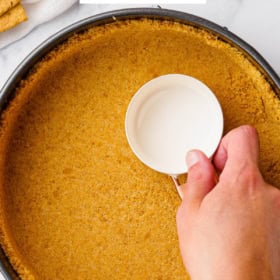 Homemade graham cracker crust in a cake pan being pressed with measuring cup.