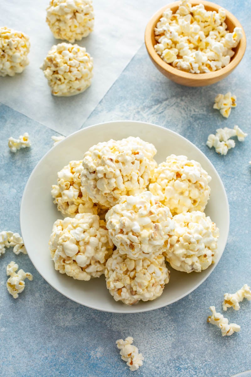 A bowl is holding several marshmallow popcorn balls with extra popcorn around the bowl.