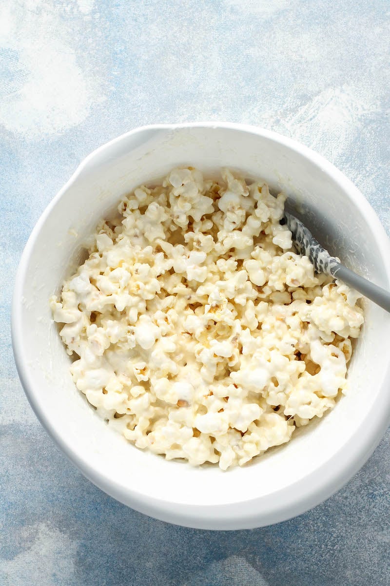 Popcorn and melted marshmallows are being stirred together in a white bowl