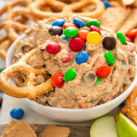 A bowl of monster cookie dough dip is on a counter with a pretzel being dipped into it