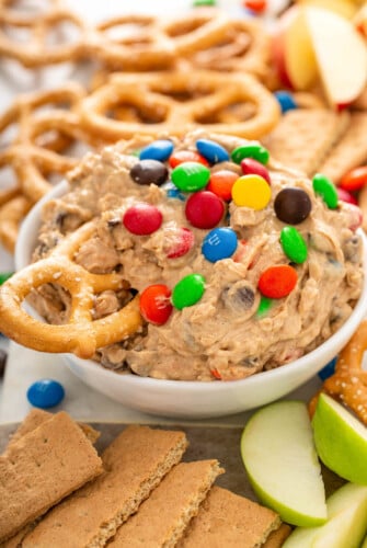A bowl of monster cookie dough dip is on a counter with a pretzel being dipped into it