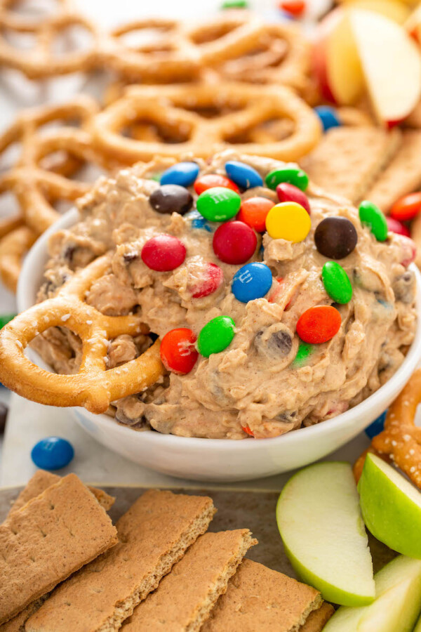 A bowl of cookie dip is on a counter with a pretzel being dipped into it