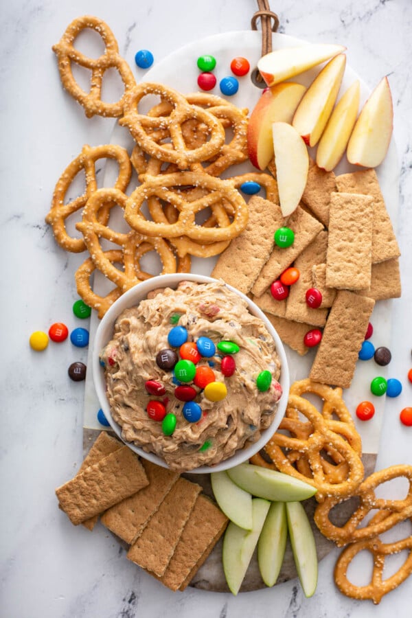 Monster cookie dough dip is on a white surface, surrounded by graham crackers and pretzels