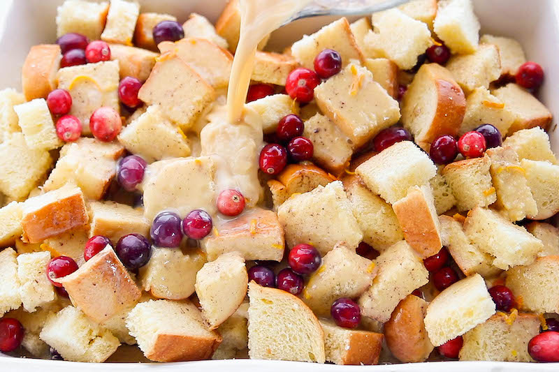 Bread and cranberries in a casserole dish with egg and milk mixture being poured on top. 