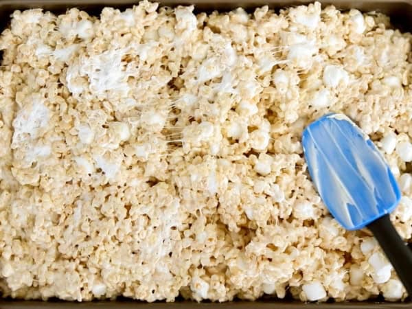 A blue rubber spatula is pressing the soft cereal and marshmallow mixture into a prepared pan.