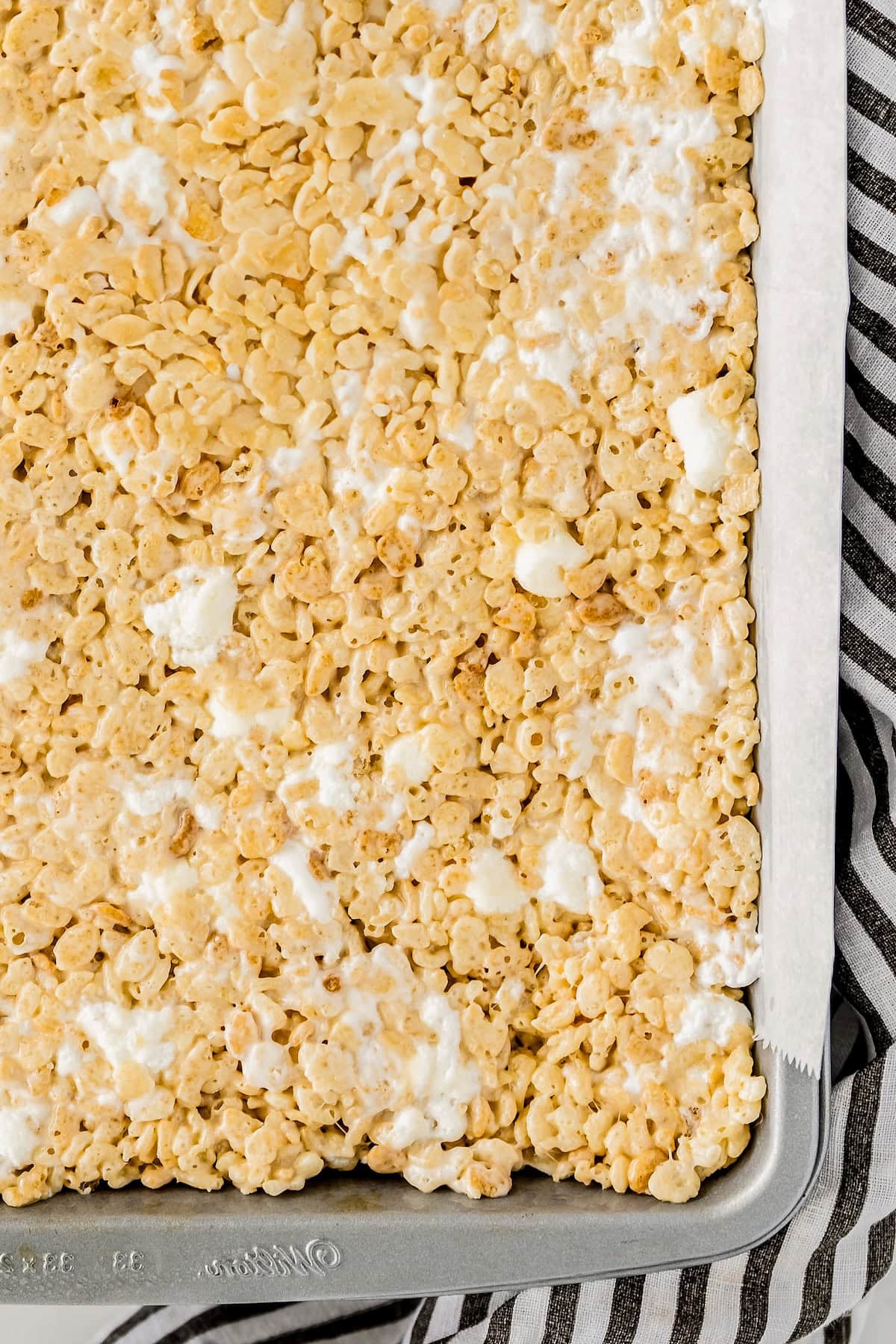 A tray filled with rice krispie treats, showing the crispy cereal bound together with melted marshmallows, resting on a sheet of parchment paper over a striped cloth.