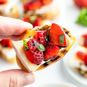 Crostini being held by a hand with strawberries, brie and balsamic vinegar drizzled on top.
