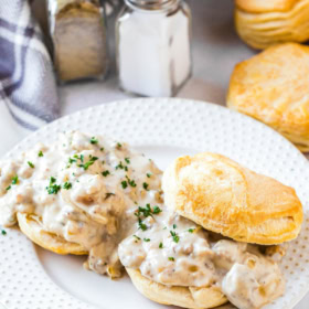 Two biscuits with sausage gravy on them on a white plate with a skillet filled with gravy in the background.